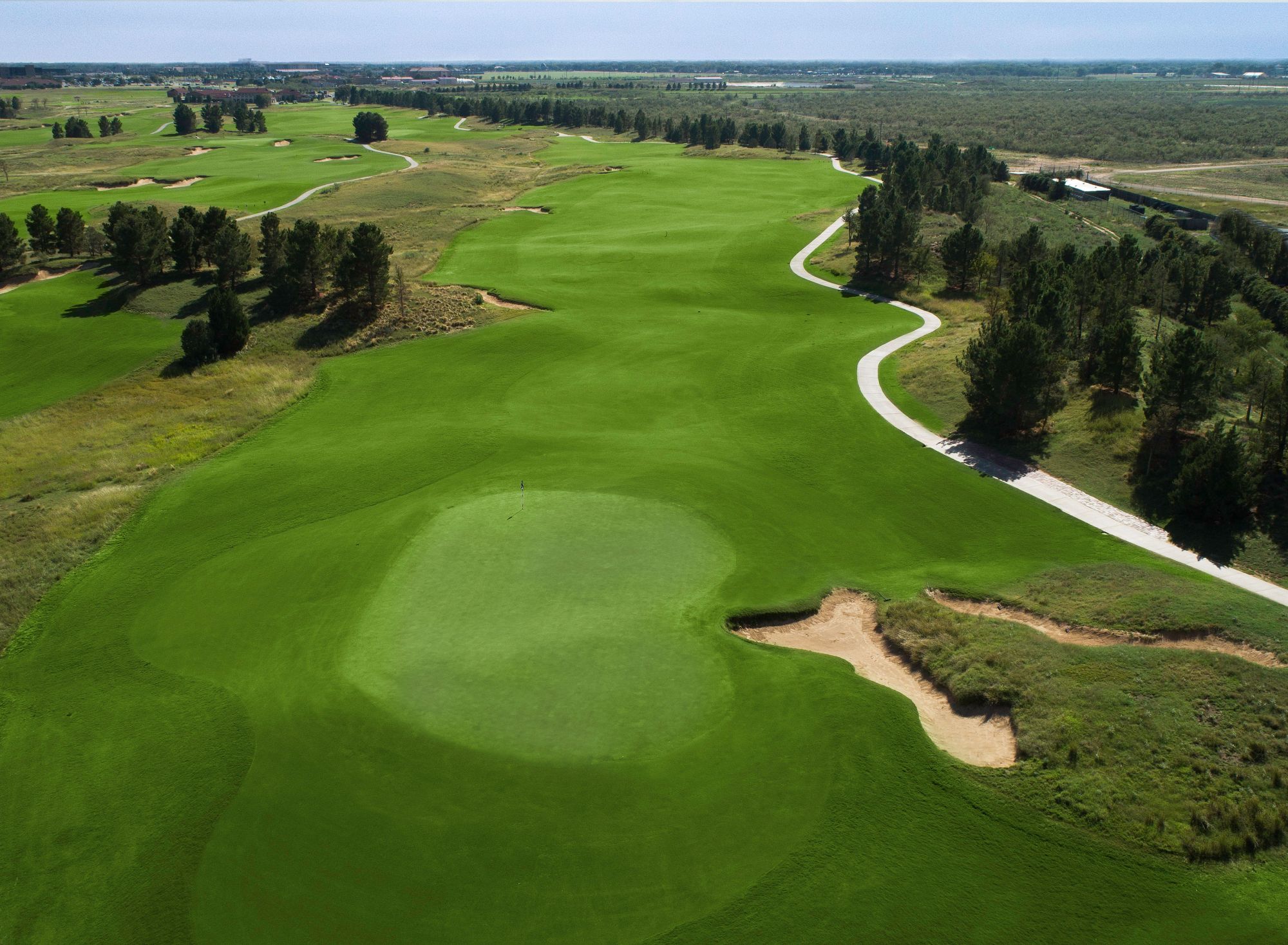 A view of a golf hole with a green fairway, trees lining the course on the right, and a clear sky overhead.